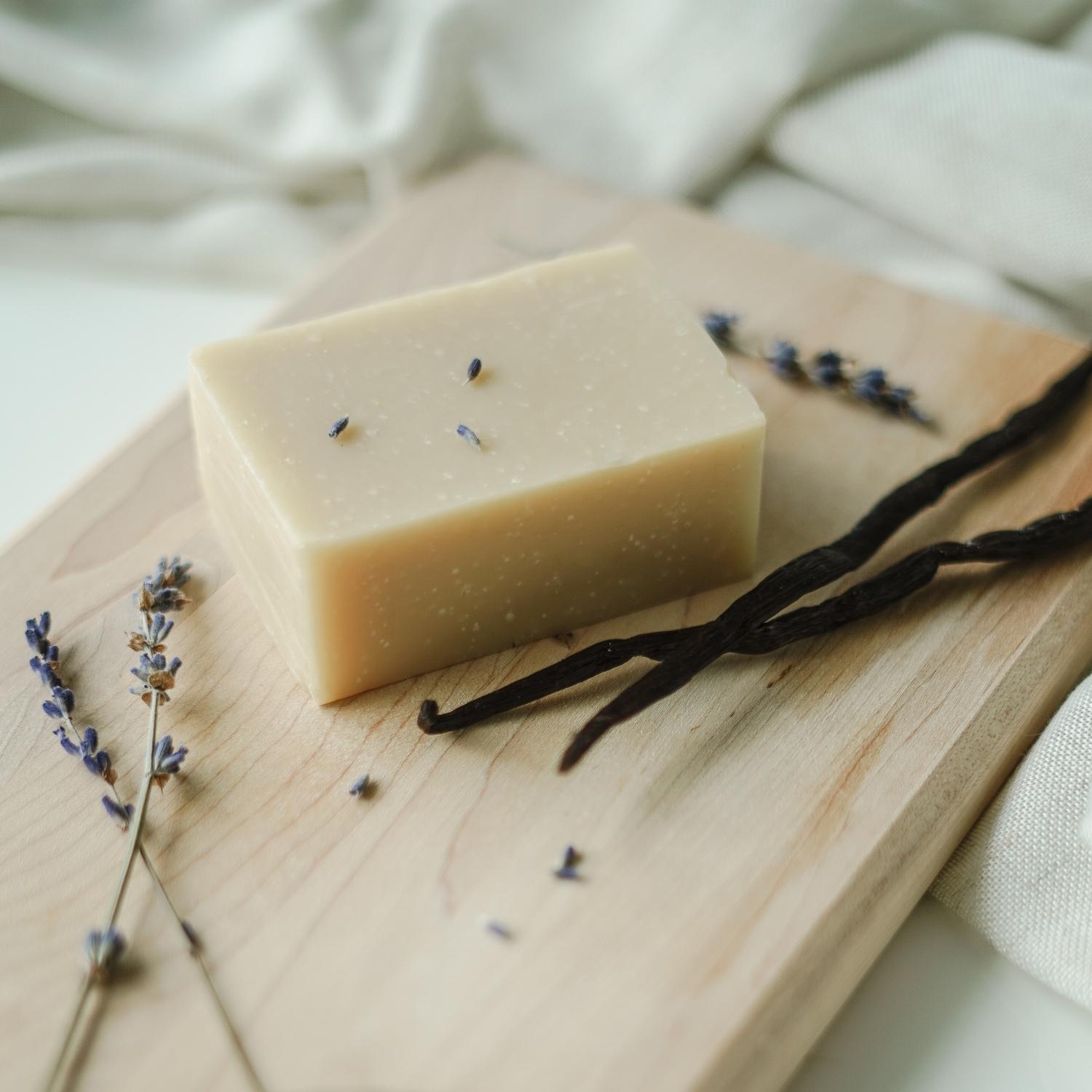 Savon en barre à la lavande et à la vanille sur une planche de bois avec des branches de lavande et de la vanille, le tout, sur un tissu blanc