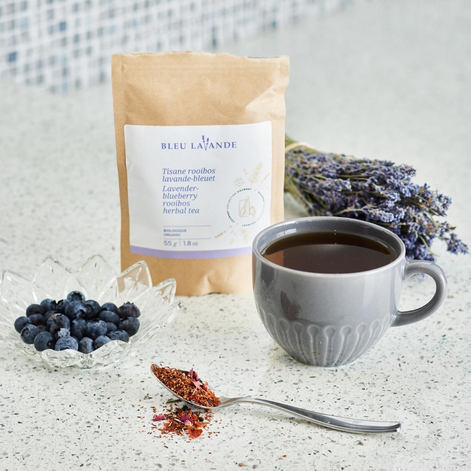 Sachet de 55g de tisane rooibos lavande-bleuet, avec une tasse de thé remplie, un pot en cristal avec des bleuets frais, une cuillère remplie de tisane et un bouquet de lavande séchée